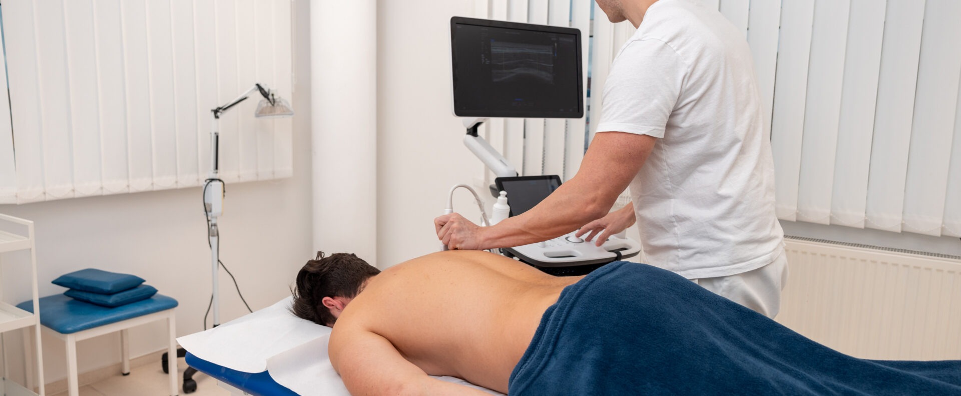 Terapeut provádí ultrazvukové vyšetření pacienta ležícího na lehátku, snímání měkkých tkání.